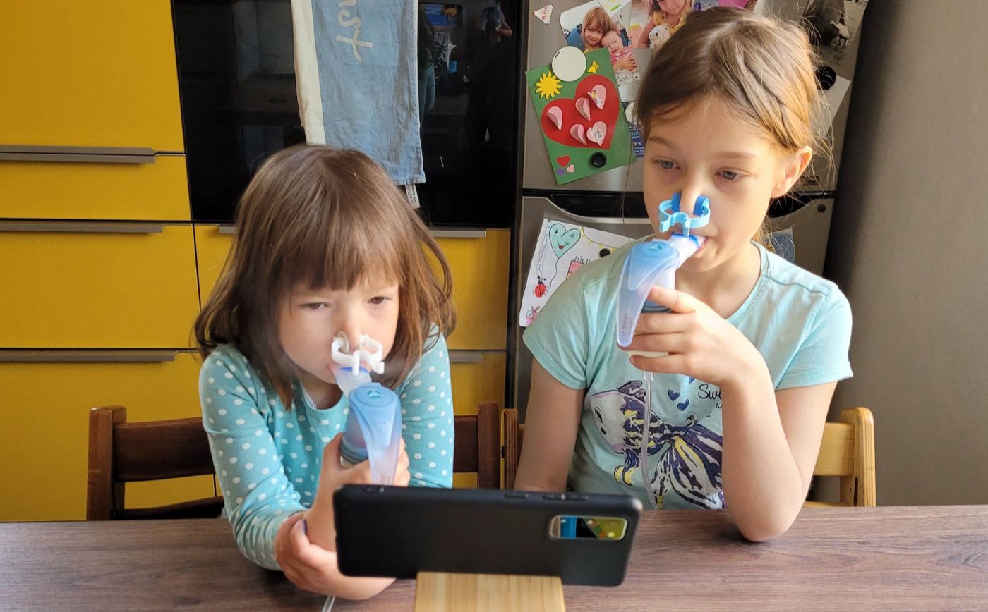 Zwei Mädchen sitzen am Tisch und inhalieren konzentriert mit Verneblern und Nasenklemmen vor einem Smartphone