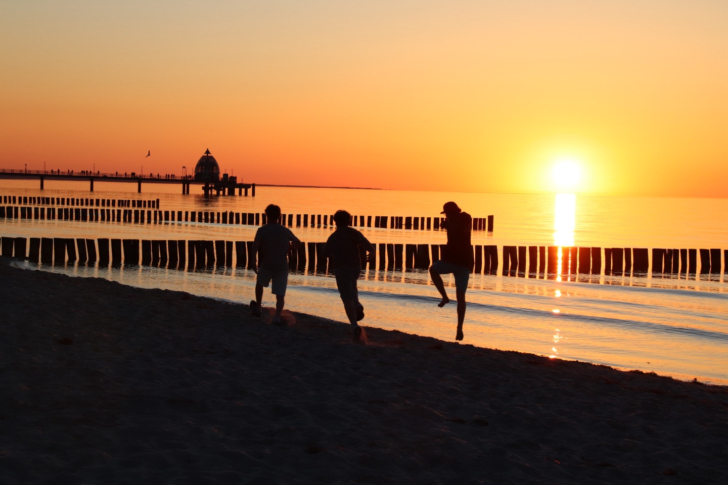 Drei Kinder springen als Silhouetten bei Sonnenuntergang an den Buhnen des Ostseestrands