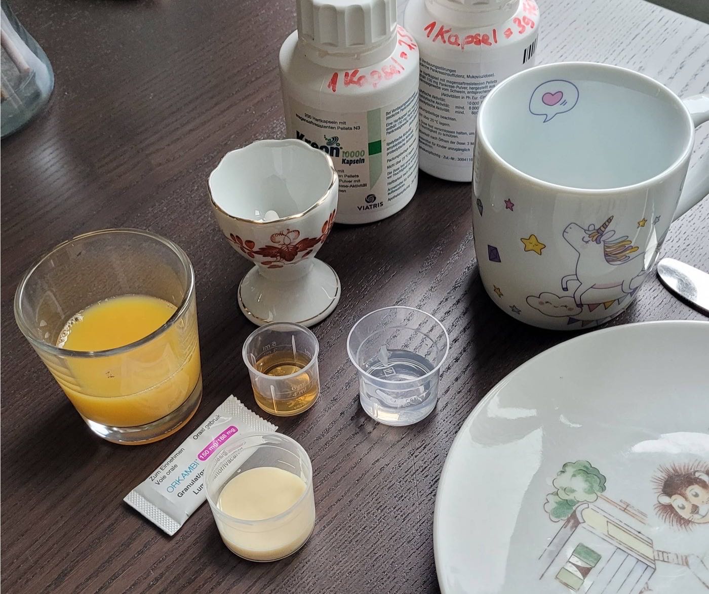 Frühstückstisch mit Medikamenten: Kreon-Kapseln, Orkambi-Granulat, Messbecher und einer Einhorntasse