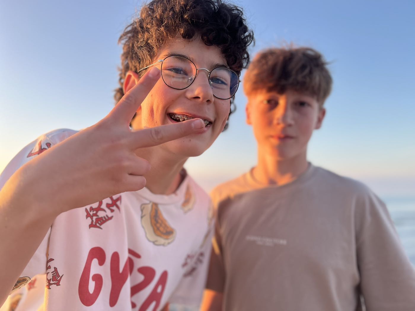 Zwei befreundete Jugendliche lachen entspannt am Meer in die Kamera, einer zeigt das Peace-Zeichen